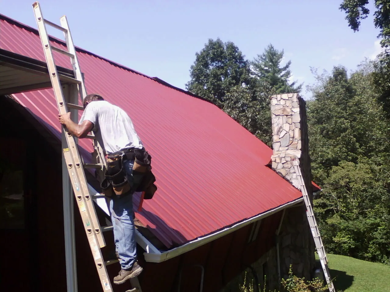 Roofer on a ladder beside a red metal roof and stone chimney — Soffit & Fascia Repair by Roofing Brothers in Forest City